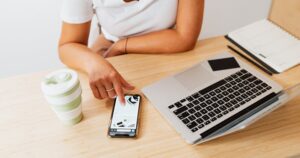 Pessoa usando smartphone ao lado de laptop em mesa de trabalho, em contexto de organização financeira e rotina digital