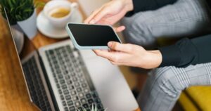 Pessoa consultando aplicativo financeiro no celular ao lado de notebook e café sobre mesa de trabalho